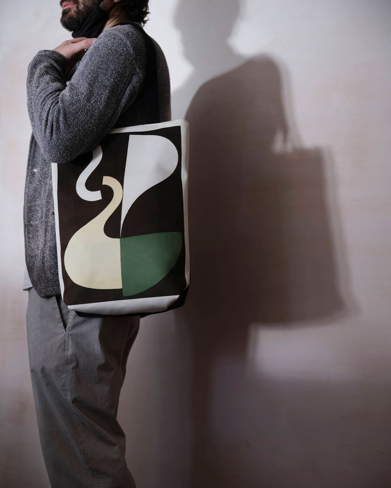 Abstract art tote bag displaying a Sophie Taeuber Arp artwork, worn by a make model throwing a shadow onto a grew wall. 