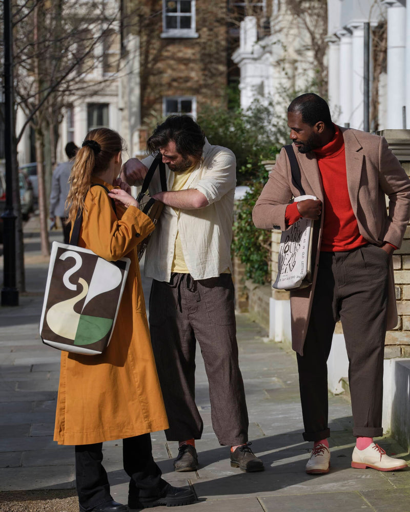 Three people on a London street wearing abstract art tote bags. One is checking his bag, the others are helping him.