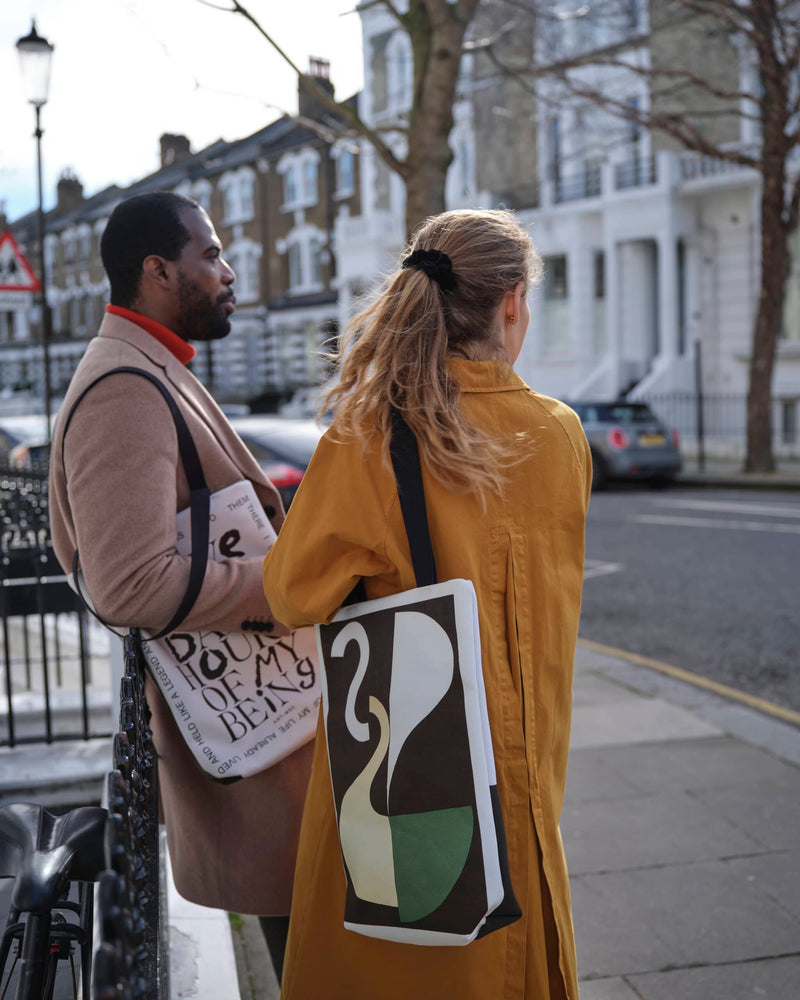 Male and female on a London street wearing abstract art tote bags. The man is viewed from the side leaning back on some black railings. The woman is in front of him with her back to us. She has long hair in a pony tail and is wearing a burnt orange trench coat.