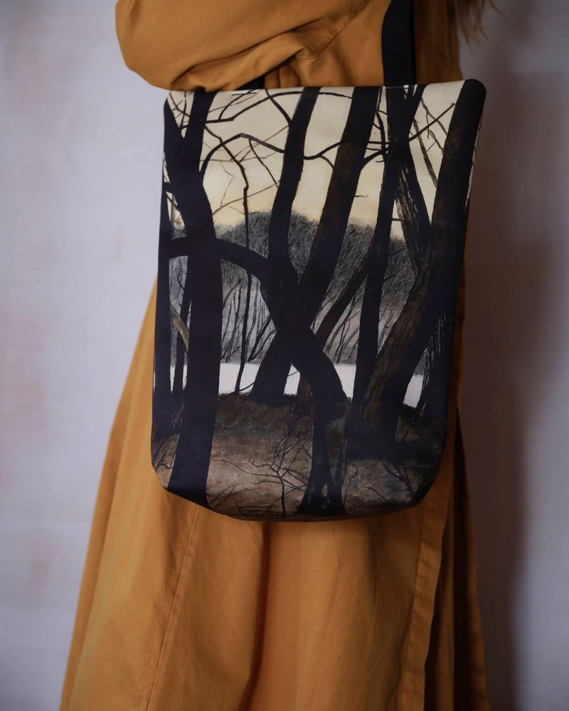 Artsy tote bag showing winter tree scene painting by Leon Spilliaert worn by female model in burnt orange trench coat, close up view.