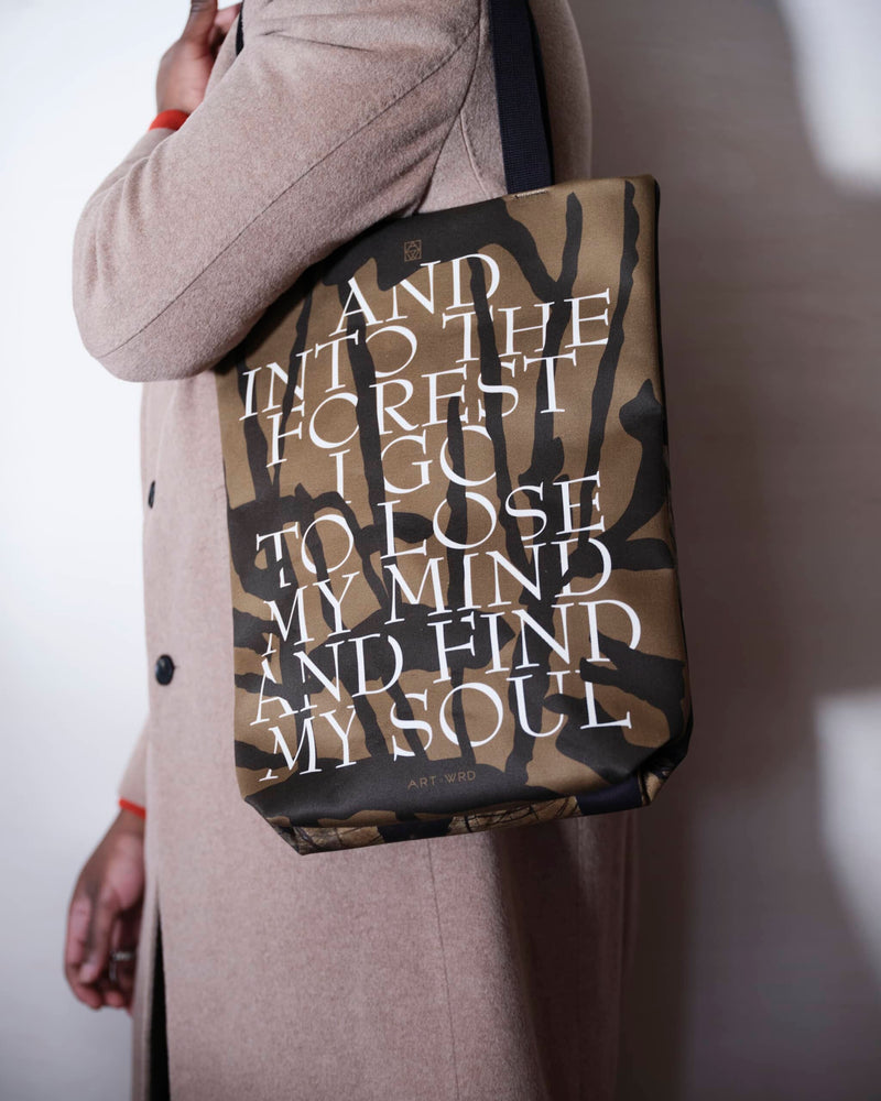 Artsy tote bag showing a forest quote worn by a male model in a light brown coat.. 
