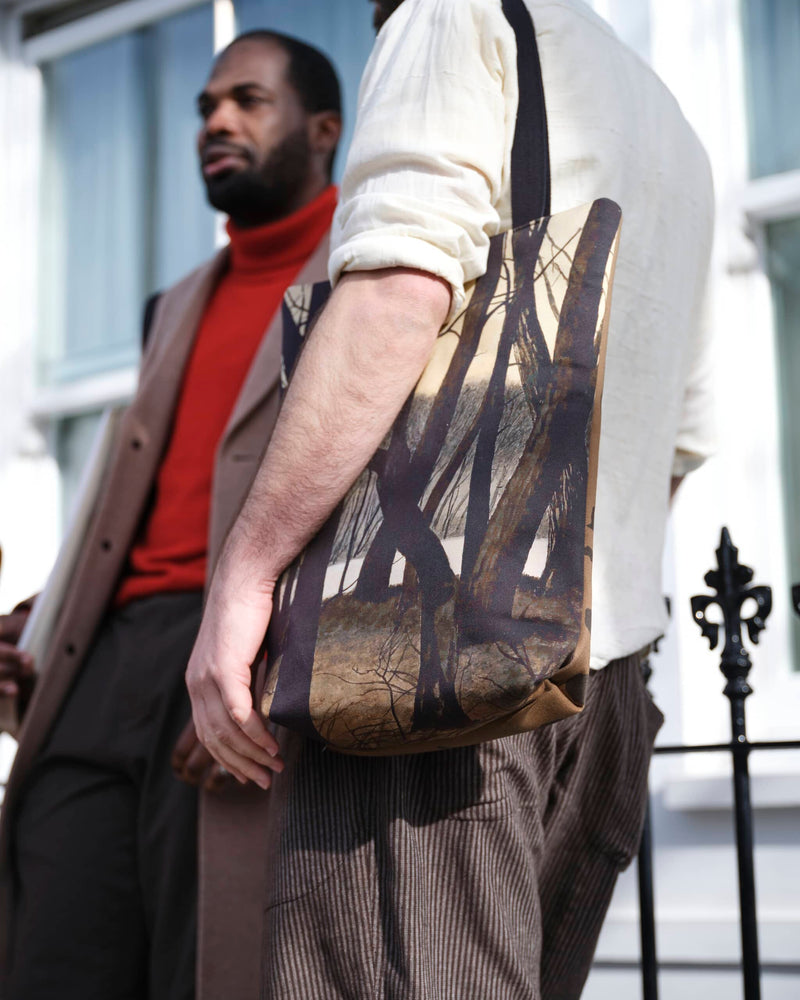 Artsy tote bag showing winter tree scene painting by Leon Spilliaert, worn by a male model stood next to black London railings. He is stood next to  another male model in a deep red roll neck leaning on the railings, looking hip.