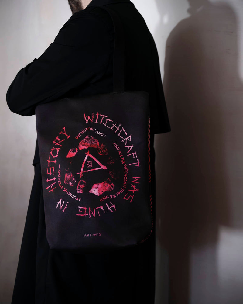 Bright pink tie-dye bag showing Emily Dickinson witches poem 'Witchcraft Was Hung', worn by a male model in a long black coat. 