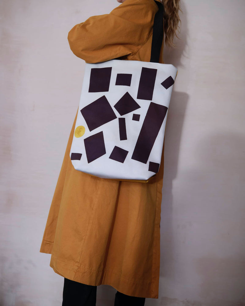 A cool, unique tote bag displaying a black and white rectangles abstract painting by Kazimir Malevich worn by a female model in a burnt orange trench coat.