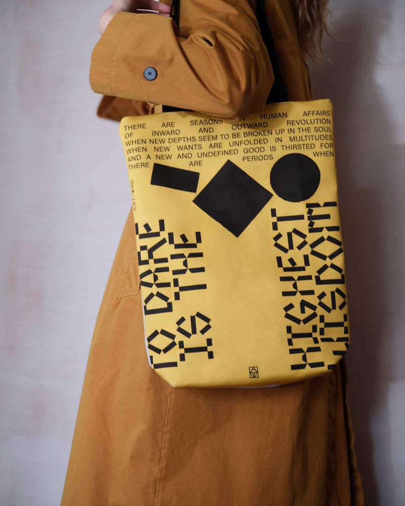 Close up of a female model wearing unique tote bag reverse of an inspiring quote presented with a futurist design. The tote bag has black writing and abstract objects on a yellow background.