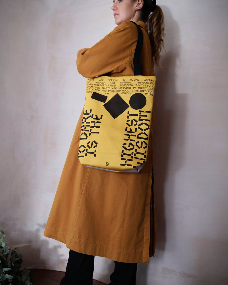 Female model wearing unique tote bag reverse of an inspiring quote presented with a futurist design. The tote bag has black writing and abstract objects on a yellow background.