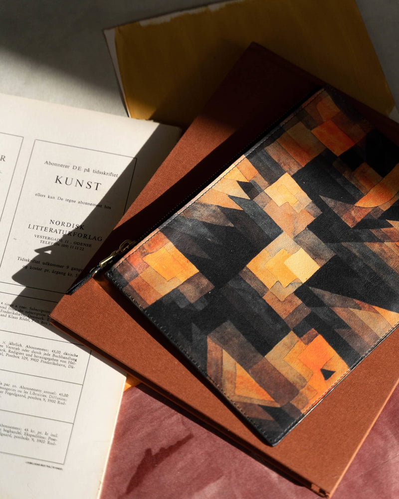 Leather art pouch printed with Paul Klee art laying on a brown fabric covered book and painted paper. The pouch is half in sunlight and half in shadow. 