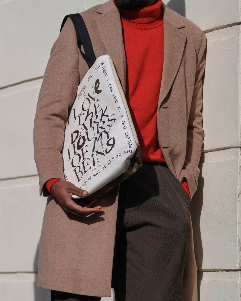Reverse of a unique art tote bag displaying a Rainer Maria Rilke quote worn by a male model. He is leaning on  a cream wall wearing an orange-red roll neck jumper and a light brown coat.