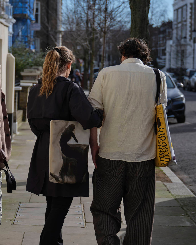 Couple viewed from behind arm in arm wearing unique art tote bags in a West London street. 